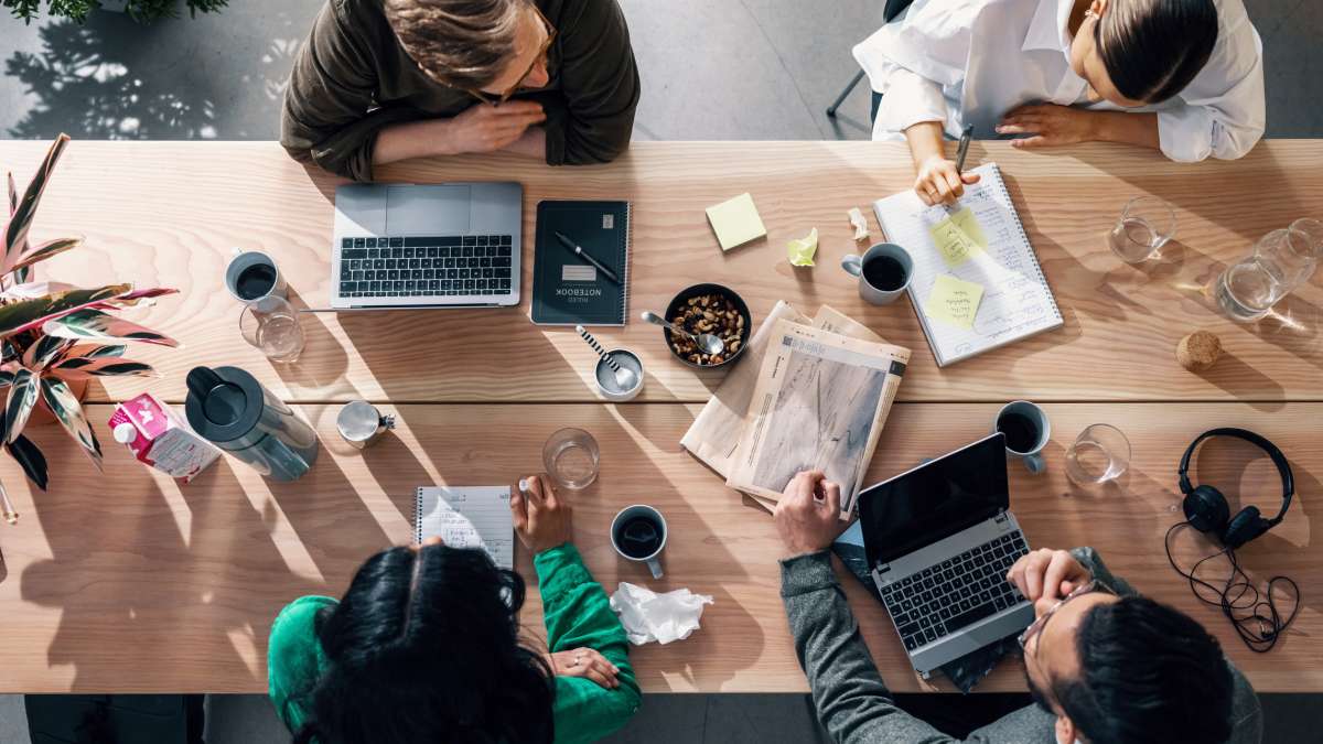 Fyra människor sitter vid ett bord, med laptops, kaffekoppar och anteckningsblock