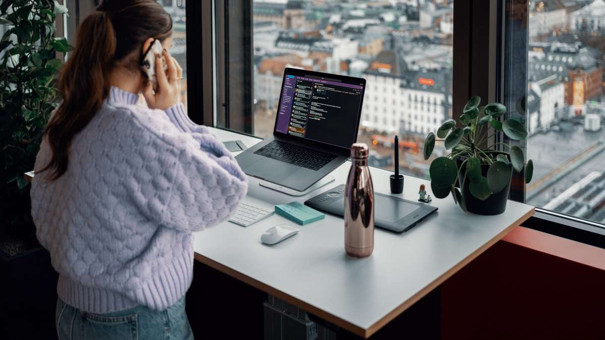 A woman is talking on a mobile phone standing in front of a laptop on a desk in front of a window